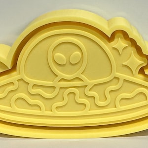 May include: A yellow, alien-themed cookie cutter. The design features a UFO with an alien and stars. The cutter is made of a solid, light yellow material and is shaped like a cloud.