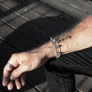 May include: A clear plastic bracelet with silver metal spikes and a silver metal buckle. The bracelet is worn on a person's wrist. The person has a tattoo on their arm that says "AFID".