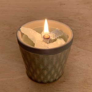 Könnte beinhalten: Eine brennende Kerze in einem hellgrünen Keramiktopf mit strukturiertem Muster. Die Kerze hat einen Holzdocht und ist von weißen Kristallen umgeben. Der Topf hat einen grauen Rand und steht auf einer Holzoberfläche.