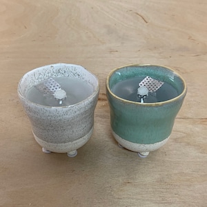 Könnte beinhalten: Zwei Kerzenhalter aus Keramik mit weißer und grüner Glasur. Jeder Halter hat einen kleinen Metall-Docht-Halter und eine weiße Kerze im Inneren.