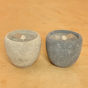 Könnte beinhalten: Zwei Beton-Kerzenhalter, einer grau und einer weiß, mit einer gesprenkelten Oberfläche. Jeder Halter hat eine kleine, runde, weiße Kerze darin.
