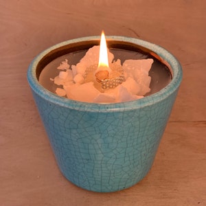 Könnte beinhalten: Ein tealfarbener Kerzenhalter aus Keramik mit einer brennenden weißen Kerze im Inneren. Die Kerze ist von weißen Wachsflocken umgeben.