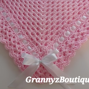 May include: A pink crocheted baby blanket with a scalloped edge and white satin ribbon accents. A white satin bow with a rhinestone center adorns the blanket. The text "GrannyzBoutique" is visible.