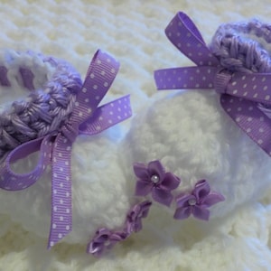 May include: A pair of white crocheted baby booties with purple crocheted trim and purple polka dot ribbon bows. The booties have purple satin flowers with pearl centers.