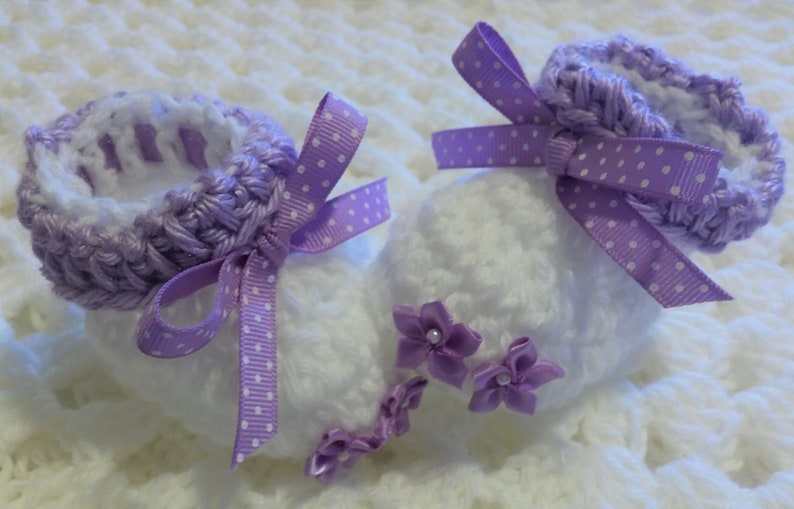 May include: A pair of white crocheted baby booties with purple crocheted trim and purple polka dot ribbon bows. The booties have purple satin flowers with pearl centers.