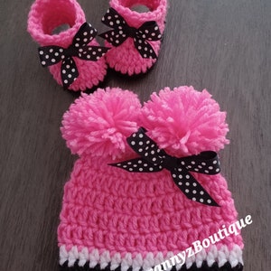 May include: Pink crocheted baby hat with black and white polka dot bow and two fluffy pom poms. Matching pink crocheted booties with black and white polka dot bows. The hat has a white and black trim. The text "GrannyzBoutique" is visible on the hat.