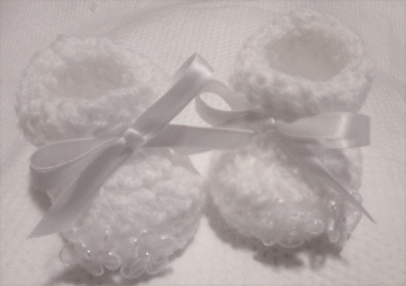 May include: Two white crocheted baby booties with white satin ribbon tied around them.