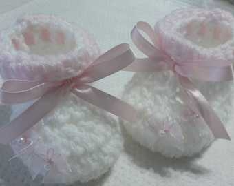 May include: A pair of white crocheted baby booties with pink satin ribbon bows and delicate lace accents.