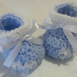 May include: A pair of baby booties knitted in a light blue color with white trim. The booties are decorated with white satin ribbon bows and white tulle bows with pearl accents.