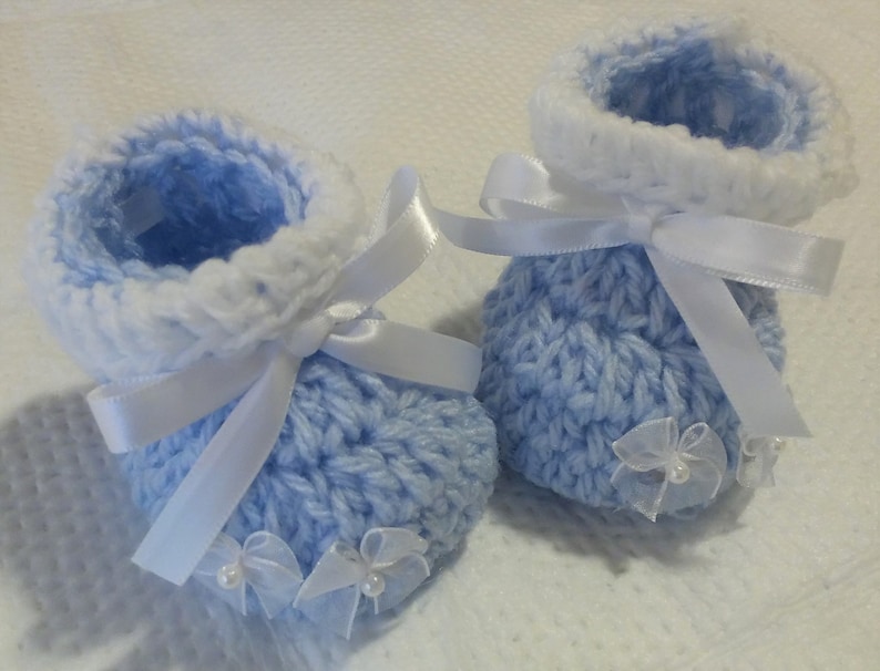 May include: A pair of baby booties knitted in a light blue color with white trim. The booties are decorated with white satin ribbon bows and white tulle bows with pearl accents.