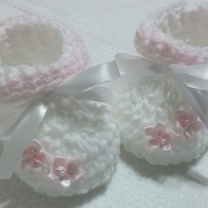 May include: Two white crocheted baby booties with pink trim and white satin ribbon bows. Each bootie has three pink satin flowers with pearl centers.