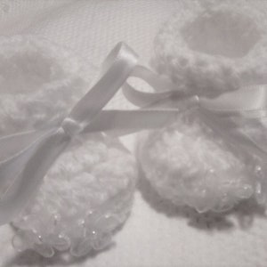 May include: Two white crocheted baby booties with white satin ribbon bows. The booties have a delicate lace trim and are perfect for a newborn.