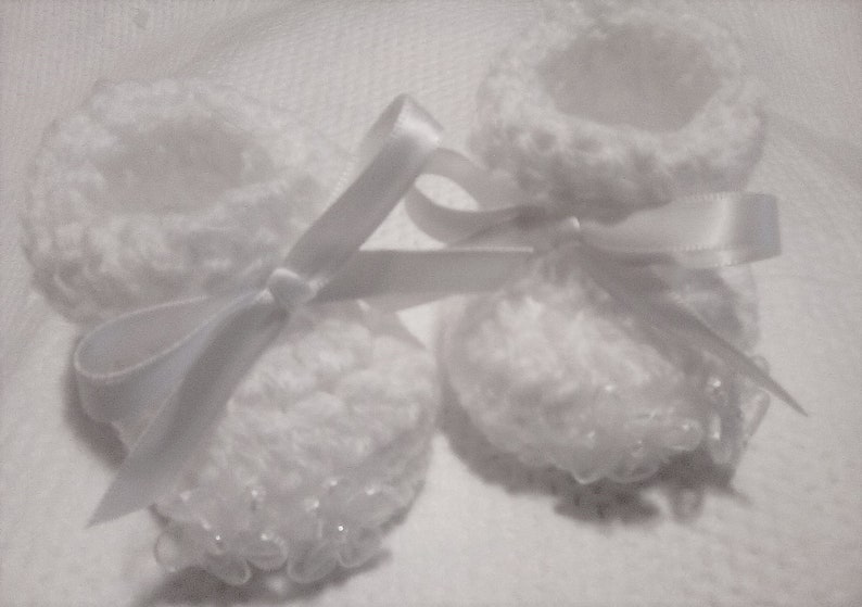 May include: Two white crocheted baby booties with white satin ribbon bows. The booties have a delicate lace trim and are perfect for a newborn.