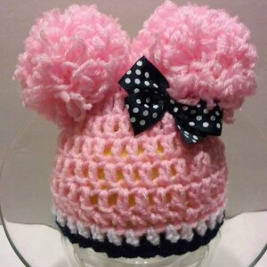 May include: Pink crocheted baby hat with two fluffy pom-poms on top and a black and white polka dot bow.