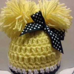 May include: Yellow crocheted hat with two yellow pom-poms on top and a black and white polka dot bow.