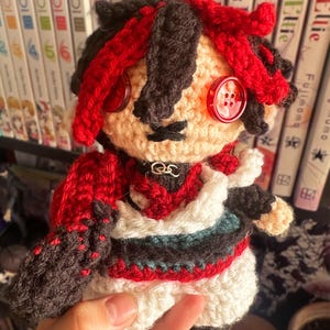 Puede incluir: Muñeca amigurumi de crochet con cabello rojo, ojos negros y un atuendo rojo, blanco y verde. La muñeca sostiene una pequeña cadena en sus manos.