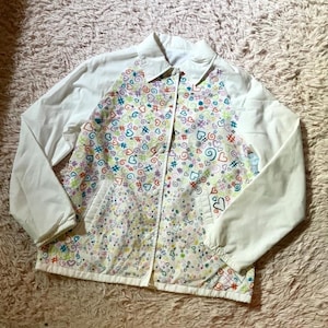May include: A white windbreaker jacket featuring a colorful all-over print on the front panels consisting of small hearts, swirls, and hashtag symbols in shades of blue, green, yellow, orange, and purple, with solid white sleeves and a collar.