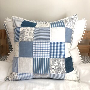 Op de afbeelding: Een blauw en wit patchwork kussen met een witte pompon rand. Het kussen is gemaakt van verschillende stoffen, waaronder denim, gingham en strepen.