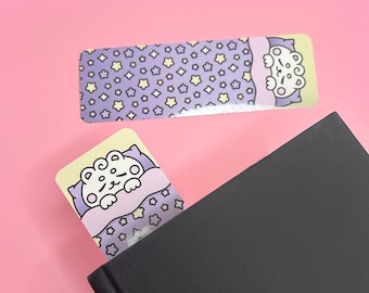 May include: A purple and yellow bookmark with a cartoon animal sleeping in a bed with stars. The bookmark is tucked into the corner of a black book.