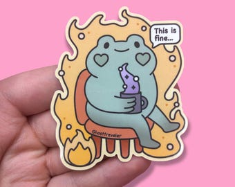 Op de afbeelding: Een sticker met een cartoonkikker met hartvormige ogen, zittend in een stoel en een mok vasthoudend. De kikker is omringd door vlammen en een tekstballon zegt: "This is fine...". De achtergrond is roze.