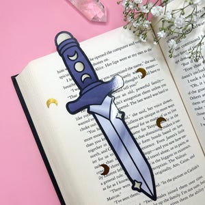 May include: A fantasy-themed bookmark shaped like a dagger, featuring a blue handle with crescent moon details and a silver blade. The bookmark is placed on an open book with text, surrounded by decorative elements.