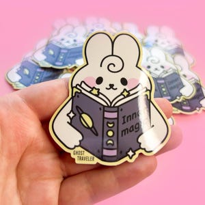 May include: A white bunny rabbit with pink cheeks is holding a purple book with yellow stars and the text "Inner Magic" on the cover. The book has a purple and yellow spine with a crescent moon and sun design. The sticker is from Ghost Traveler.