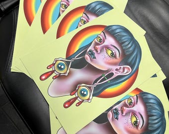 May include: A stack of art prints featuring a stylized illustration of a woman with blue hair, yellow eyes, and a black lip. The woman has an eye-shaped earring with a red teardrop. A rainbow arc is behind the woman's head.