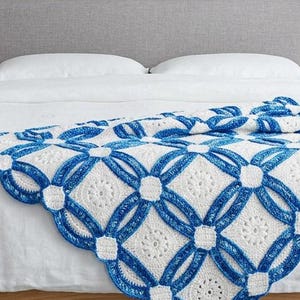 May include: A white and blue crochet blanket with a geometric design is draped over a bed. The blanket features a repeating pattern of interlocking circles and squares in white and blue. The bed has a light wood frame and a gray headboard.