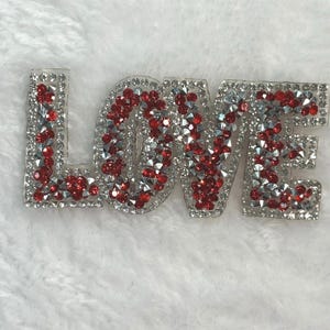 May include: A clear, rhinestone-studded "LOVE" word art piece. The letters are outlined with clear rhinestones and filled with a mix of red and silver rhinestones, creating a sparkling effect. The piece is set against a white, textured background.