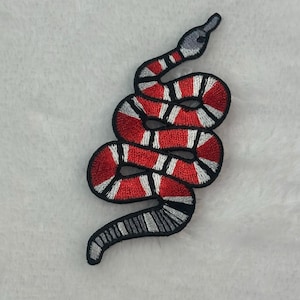 May include: Embroidered snake patch featuring a red and white pattern on a black outline. The snake's head is gray, and the body is coiled. This patch is suitable for clothing and accessories.