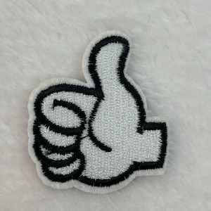 May include: Embroidered patch in the shape of a thumbs-up hand gesture. The patch features a white hand with black outlining and a black border. The patch is on a white, textured background.