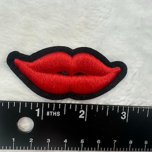 May include: Embroidered red lips patch with a black border. The patch is approximately 2 inches wide. The lips are a bright red color, and the black border provides a defined outline. The patch is on a white, textured surface.