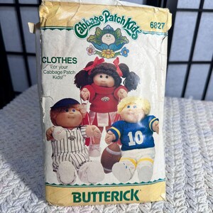 May include: Vintage Butterick sewing pattern for Cabbage Patch Kids clothes. The package features illustrations of Cabbage Patch Kids dolls in various outfits, including a cheerleader, baseball player, and football player. The package is yellowed with age.