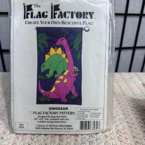 May include: A packaged "Flag Factory" pattern for a dinosaur-themed flag. The flag design features a purple and green dinosaur on a dark blue background. The package includes instructions for a flag that finishes 28" x 49" and is easy to sew.