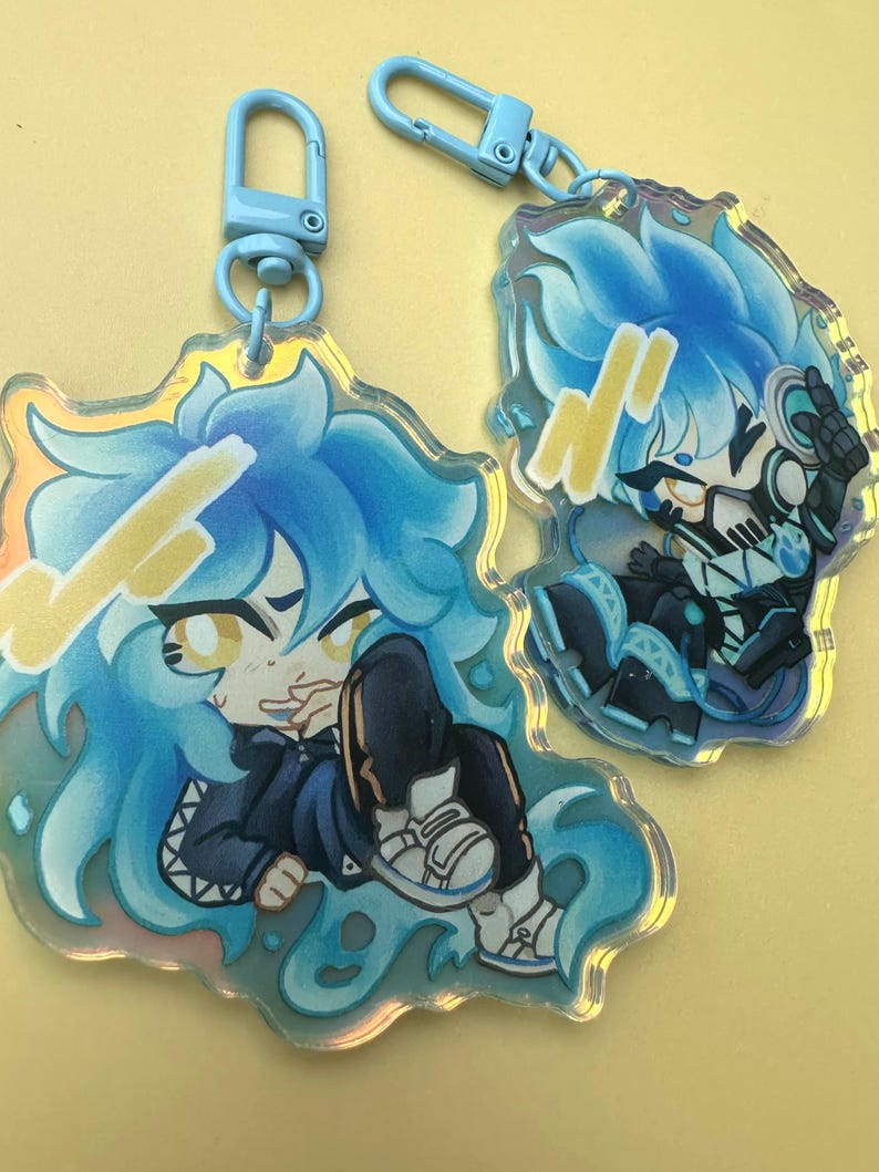 Puede incluir: Dos llaveros acr&iacute;licos con personajes de estilo anime con cabello y atuendos azules. Los llaveros tienen un efecto hologr&aacute;fico y est&aacute;n unidos a cierres azul claro. Un personaje est&aacute; sentado, el otro en una pose din&aacute;mica.