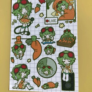 May include: A sticker sheet featuring a grid background with multiple illustrations of a green-haired character wearing red goggles and an orange outfit. The sheet includes various poses, carrots, a crate of carrots, and a box labelled OLIVE.