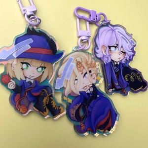 Puede incluir: Tres llaveros acr&iacute;licos con personajes de estilo anime. Cada llavero presenta un dise&ntilde;o de personaje diferente, con diferentes colores de cabello y atuendos. Los llaveros est&aacute;n sujetos a cierres met&aacute;licos de color p&uacute;rpura. El fondo es de color amarillo claro liso.