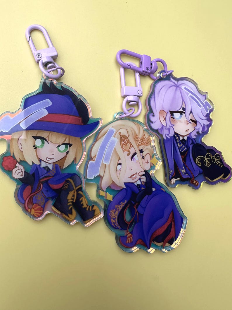 Puede incluir: Tres llaveros acr&iacute;licos con personajes de estilo anime. Cada llavero presenta un dise&ntilde;o de personaje diferente, con diferentes colores de cabello y atuendos. Los llaveros est&aacute;n sujetos a cierres met&aacute;licos de color p&uacute;rpura. El fondo es de color amarillo claro liso.