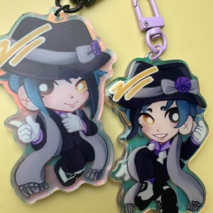 Puede incluir: Dos llaveros de personajes de dibujos animados. Cada llavero presenta un personaje con sombrero, traje y bufanda. Los llaveros tienen un efecto hologr&aacute;fico y est&aacute;n sujetos a un cierre negro y morado.