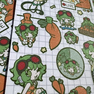 May include: A glittery sticker sheet featuring multiple cartoon illustrations of a green-haired character wearing red goggles and an orange outfit. The designs include the character with carrots, a live stream frame, and various poses on a grid background.