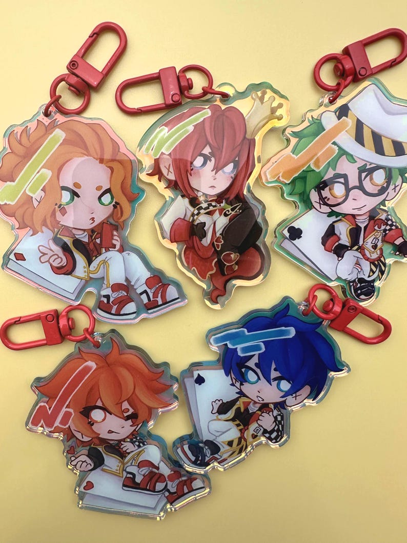Puede incluir: Llaveros acr&iacute;licos con personajes de estilo anime con cierres rojos de pinza de langosta. Los llaveros muestran personajes con varios colores de cabello, atuendos y motivos de naipes. Cada personaje est&aacute; representado en un estilo chibi, con colores vibrantes y dise&ntilde;os detallados.