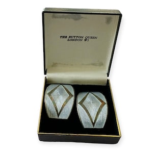 May include: A pair of silver-toned cufflinks with a geometric design, presented in a black box with gold trim. The cufflinks feature a diamond-shaped center with radiating lines. The box is labeled "THE BUTTON QUEEN LONDON W1".