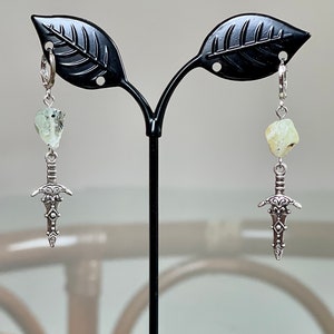 Op de afbeelding: Een paar zilveren dolk-oorbellen met een lichtgroene stenen hanger die aan elke oorbel hangt. De oorbellen worden getoond op een zwarte oorbelstandaard.