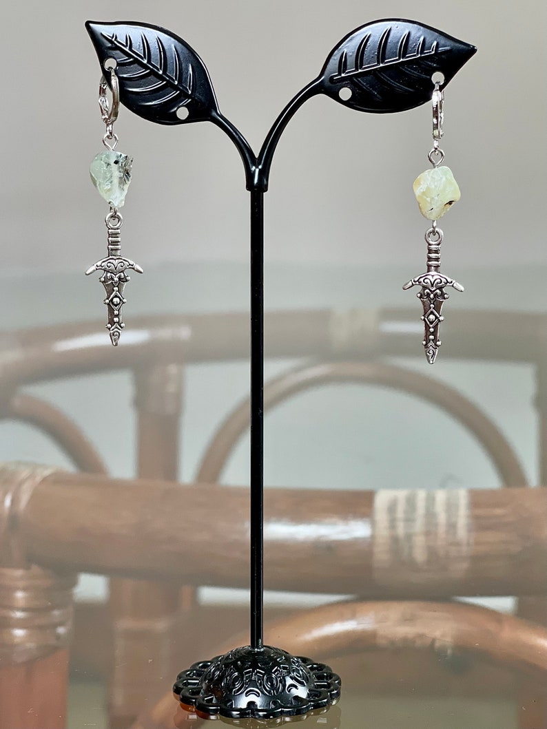 Op de afbeelding: Een paar zilveren dolk-oorbellen met een lichtgroene stenen hanger die aan elke oorbel hangt. De oorbellen worden getoond op een zwarte oorbelstandaard.