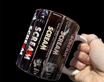 Peut inclure: Une tasse en céramique blanche avec un motif de quatre cassettes VHS empilées les unes sur les autres. Les cassettes sont étiquetées "SCREAM" et présentent une image en noir et blanc d'un visage de femme sur chaque cassette.