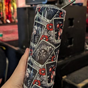May include: A stainless steel tumbler featuring a black and white tarot card design. The design includes images of a skeleton, a heart, and red flowers. The words "THE FOOL" and "THE LOVERS" are visible on the tumbler.