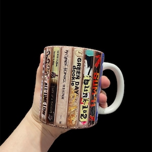 May include: A white ceramic mug with a design of stacked cassette tapes featuring the band names Blink-182, Green Day, Dookie, My Chemical Romance, Too Weird To Live, Too Rare To Die, Death of a Bachelor, and All Time Low.