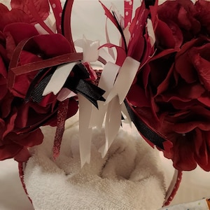 May include: Red fabric flower headband with black, white, and red ribbon accents. The headband is on a white fluffy surface.