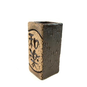 May include: A rectangular ceramic vase with a textured, dark brown exterior and a tan front panel. The front panel features black Japanese characters within a black oval frame. The vase has a square opening at the top.