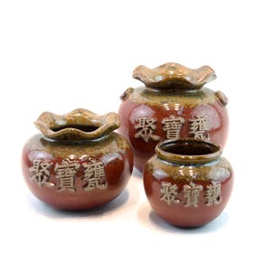 May include: Three ceramic jars in varying sizes, each with a reddish-brown base and a ruffled, light brown rim. The jars have Chinese characters embossed on their surface. The jars are likely decorative or storage containers.